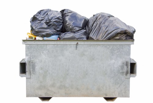 A large industrial skip positioned outdoors on a paved surface, filled with multiple tightly packed black rubbish bags of varying sizes. Some of the bags appear to be made of plastic with a slightly reflective surface, while others are matte, indicating different materials. The skip is constructed from galvanized steel with a weathered, metallic finish, and has prominent handles on each side for lifting. The bags are stacked unevenly, with some leaning against each other, and extend slightly above the top edge of the skip, suggesting recent or ongoing disposal activity. The background shows an outdoor setting, potentially near a commercial or industrial area, with diffuse lighting indicating an overcast sky. This scene is representative of waste removal operations conducted by companies such as Commercial Waste Removal Brimsdown, servicing local businesses in the Enfield area within the postal code region relevant to Brimsdown and surrounding locations. The image emphasizes the type of refuse typically handled in commercial rubbish collection services, highlighting the scale and material diversity of waste managed in such operations.